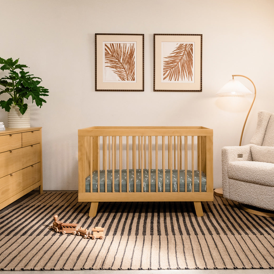 Front view of Babyletto Hudson 3-in-1 Convertible Crib And Toddler Rail next to dresser and recliner in -- Color_Honey